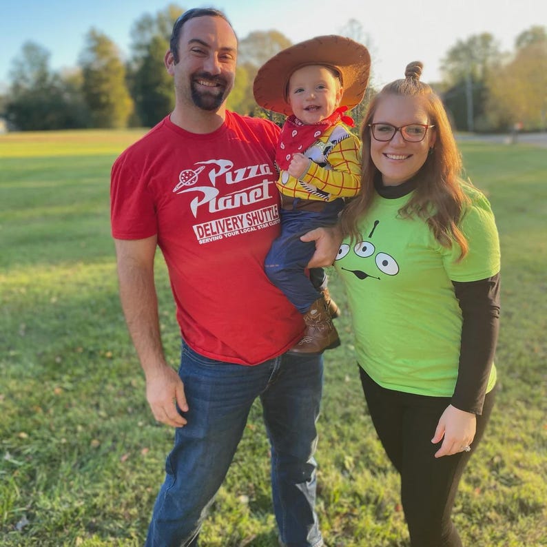 May include: A family dressed in costumes poses outdoors. The father wears a red Pizza Planet t-shirt, the child is dressed as Woody from Toy Story, and the mother wears a green shirt with an alien graphic. The background is a grassy field.
