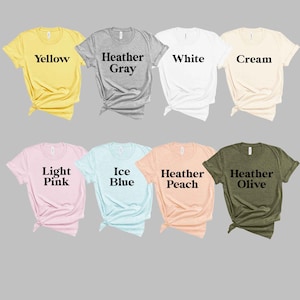 以下が含まれることがあります： 黄色、ヘザーグレー、白、クリーム、ライトピンク、アイスブルー、ヘザーピーチ、ヘザーオリーブなど、さまざまな色のユニセックスTシャツのコレクション。Tシャツはすべて折り畳んでグレーの背景に並べてあります。