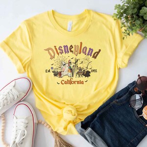 May include: Yellow t-shirt with a graphic of the Nightmare Before Christmas characters in front of a Disneyland sign that says "Disneyland Est. 1955 California".