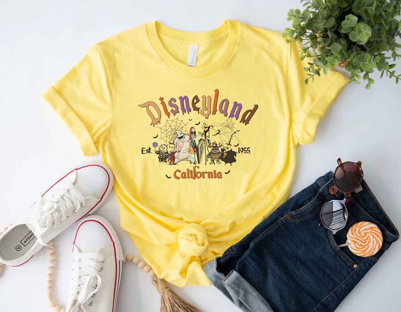 May include: Yellow t-shirt with a graphic of the Nightmare Before Christmas characters in front of a Disneyland sign that says "Disneyland Est. 1955 California".