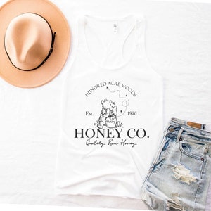 May include: White tank top with a black and white graphic of Winnie the Pooh holding a honey pot. The graphic also includes the text "Hundred Acre Woods", "Est. 1926", "Honey Co.", and "Quality Raw Honey".