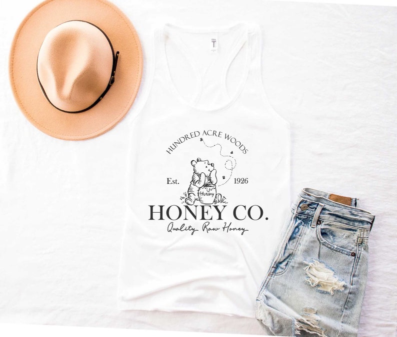May include: White tank top with a black and white graphic of Winnie the Pooh holding a honey pot. The graphic also includes the text "Hundred Acre Woods", "Est. 1926", "Honey Co.", and "Quality Raw Honey".