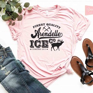 Puede incluir: Una camiseta rosa con texto negro que dice "Finest Quality Arendelle Ice Co. Established 2013". El texto est&aacute; en una fuente de estilo vintage e incluye un gr&aacute;fico de dos picos cruzados y una silueta de un alce.