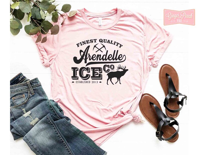 Puede incluir: Una camiseta rosa con texto negro que dice "Finest Quality Arendelle Ice Co. Established 2013". El texto est&aacute; en una fuente de estilo vintage e incluye un gr&aacute;fico de dos picos cruzados y una silueta de un alce.