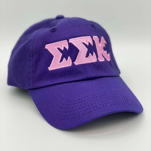 May include: A purple baseball cap with a curved brim. The cap features the embroidered Greek letters ΣΣΚ in light pink on the front. The hat is made of a durable fabric and has a classic, adjustable design.