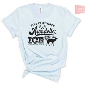 Puede incluir: Camiseta azul claro con texto negro que dice "Finest Quality Arendelle Ice Co. Established 2013". El texto est&aacute; en una fuente de estilo vintage e incluye un gr&aacute;fico de dos picos cruzados y una silueta de un reno.