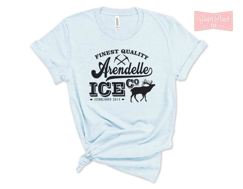 Puede incluir: Camiseta azul claro con texto negro que dice "Finest Quality Arendelle Ice Co. Established 2013". El texto est&aacute; en una fuente de estilo vintage e incluye un gr&aacute;fico de dos picos cruzados y una silueta de un reno.