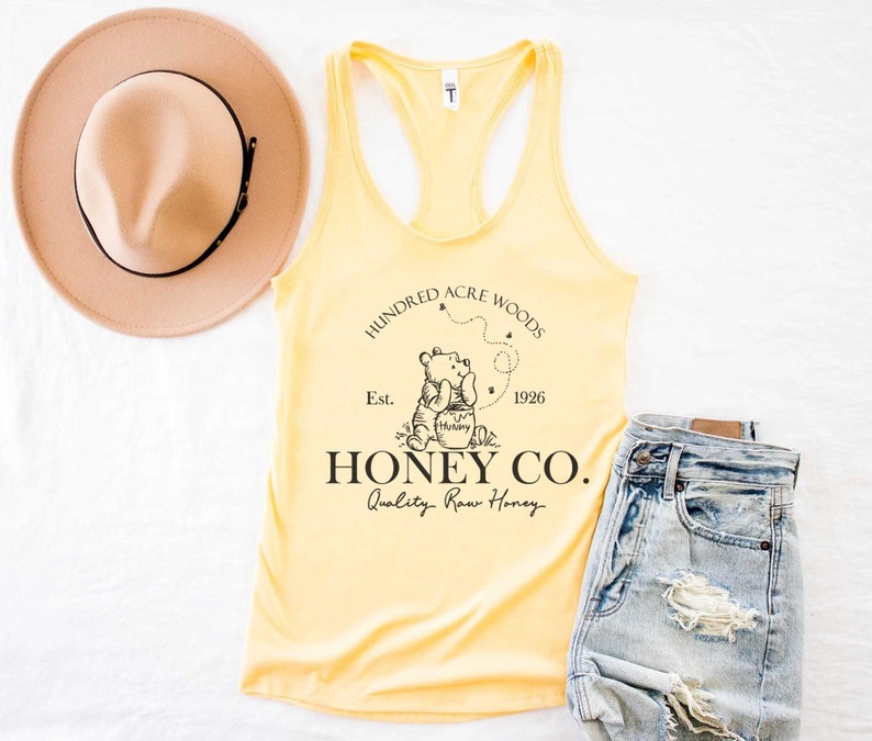 May include: Yellow tank top with a black graphic of Winnie the Pooh and a honey pot. The graphic includes the text "Hundred Acre Woods", "Est. 1926", and "Honey Co. Quality Raw Honey".