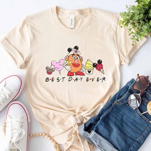 May include: A beige t-shirt with a graphic of a cartoon character wearing a nurse's hat and holding a Mickey Mouse ice cream cone. The text "BEST DAY EVER" is printed below the graphic.