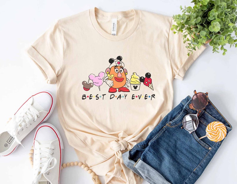 May include: A beige t-shirt with a graphic of a cartoon character wearing a nurse's hat and holding a Mickey Mouse ice cream cone. The text "BEST DAY EVER" is printed below the graphic.