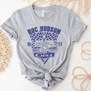 以下が含まれることがあります： 映画「カーズ」のキャラクター、ドック・ハドソンが描かれたグレーのTシャツ。デザインには「Doc Hudson」、「19」、「51」、「Cars」の文字が書かれています。