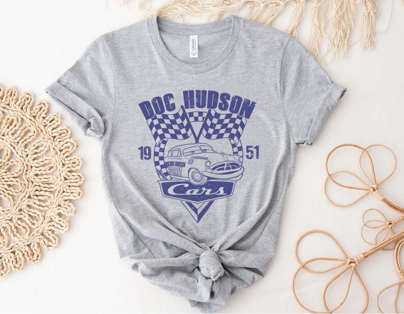 以下が含まれることがあります： 映画「カーズ」のキャラクター、ドック・ハドソンが描かれたグレーのTシャツ。デザインには「Doc Hudson」、「19」、「51」、「Cars」の文字が書かれています。