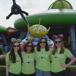 May include: Four people wearing lime green t-shirts with a three-eyed alien face graphic. They are posing in front of a large green alien statue and a spaceship-themed ride. Some are wearing sunglasses and mouse ear headbands.