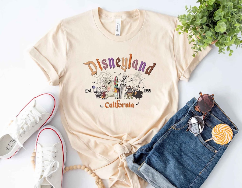 May include: A beige t-shirt with a graphic print of the Disneyland logo and characters from the Nightmare Before Christmas. The text reads "Disneyland Est. 1955 California".