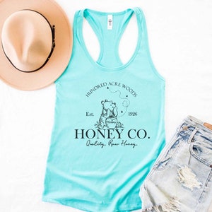 May include: A turquoise tank top with a black graphic of Winnie the Pooh holding a honey pot. The graphic also includes the text "Hundred Acre Woods", "Est. 1926", "Honey Co.", and "Quality Raw Honey".
