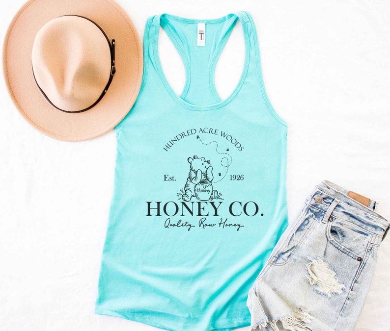 May include: A turquoise tank top with a black graphic of Winnie the Pooh holding a honey pot. The graphic also includes the text "Hundred Acre Woods", "Est. 1926", "Honey Co.", and "Quality Raw Honey".