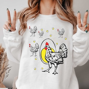 May include: White sweatshirt with a graphic of a chicken inside an egg and other chickens around it. The chickens are black and white with red combs.