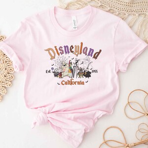 May include: Pink t-shirt with a graphic print of the Nightmare Before Christmas characters in front of a Disneyland logo. The text reads "Disneyland Est. 1955 California".