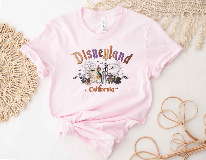 May include: Pink t-shirt with a graphic print of the Nightmare Before Christmas characters in front of a Disneyland logo. The text reads "Disneyland Est. 1955 California".