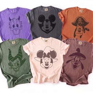 May include: Six short-sleeved crew neck t-shirts in various colors including purple, charcoal, orange, olive green, light pink, and maroon. Each shirt features a different screen-printed character graphic in a dark ink color on the front chest area.