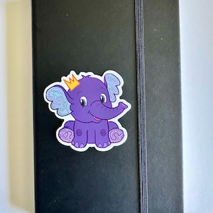 May include: A black notebook with an elastic closure and a purple elephant sticker. The sticker features a cartoon elephant with a gold crown, blue patterned ears, and purple feet. The notebook is closed.