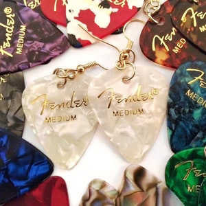 May include: A pair of gold earrings with white, pearlescent guitar picks that say "Fender Medium" in gold lettering.