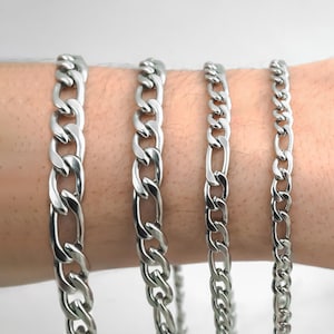 Peut inclure: Quatre bracelets à chaîne figaro en métal argenté de différentes épaisseurs disposés côte à côte. Chaque bracelet présente un motif répétitif de trois petits maillons circulaires suivis d'un maillon ovale allongé, avec une finition polie.