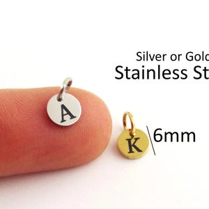 Könnte beinhalten: Zwei kleine kreisförmige Anhänger aus Edelstahl, einer in Silberoptik mit dem Buchstaben A und einer in Goldoptik mit dem Buchstaben K. Jeder Anhänger hat einen Biegering und misst 6mm im Durchmesser. Der Text Silver or Gold Stainless Steel ist sichtbar.