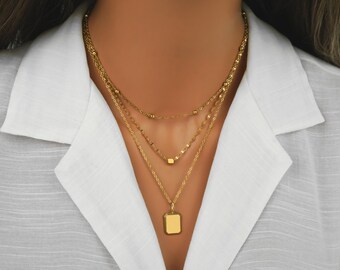 May include: A gold-toned layered necklace featuring three distinct chains. The top chain has small beads, the middle chain includes a small square charm, and the bottom chain holds a rectangular pendant with a textured border and a smooth center.