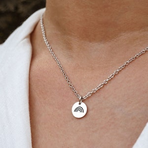 May include: A silver-colored necklace with a small, round pendant. The pendant features a black rainbow design. The necklace is worn against a white shirt.