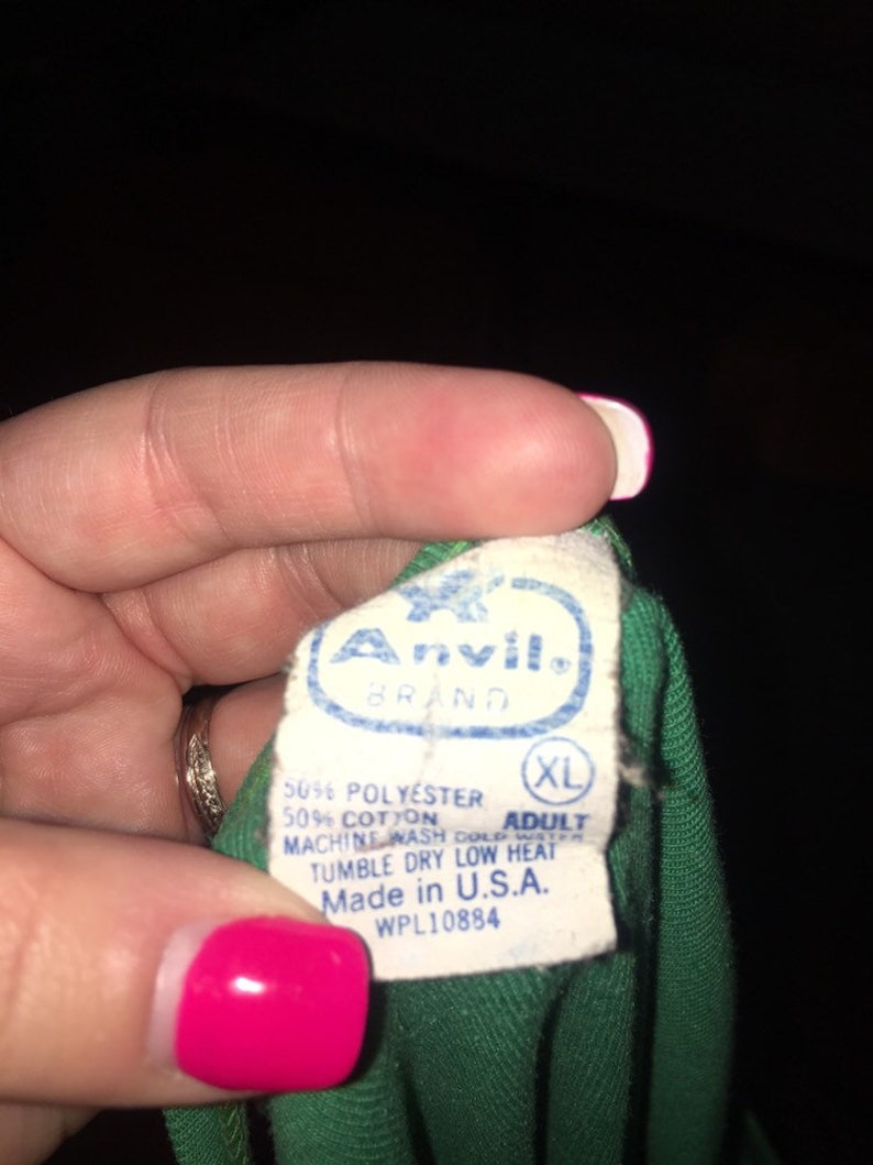 May include: A green t-shirt tag with the Anvil brand name and logo. The tag also includes the size XL, fabric content, care instructions, and "Made in U.S.A." with the WPL number 10884.