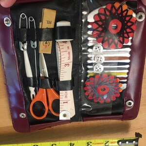 May include: A burgundy leather sewing kit with a clear plastic window. The kit contains a variety of sewing supplies, including needles, thread, buttons, a thimble, and a small pair of scissors. The kit also includes a small tape measure that reads "1 inch" and "3 Made in 4".