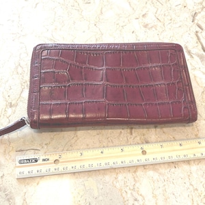 May include: A burgundy-colored wallet with a crocodile skin texture. The wallet has a zipper closure and a small pull tab. A ruler is placed next to the wallet for scale.