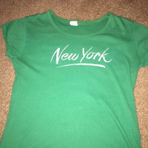 May include: Green short-sleeved t-shirt with white text that says "New York" in a cursive font.