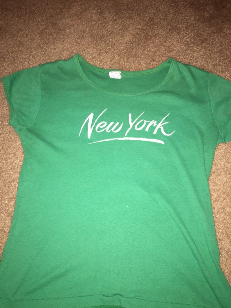 May include: Green short-sleeved t-shirt with white text that says "New York" in a cursive font.