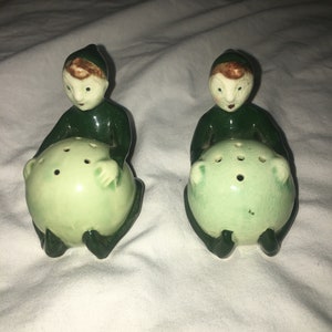 May include: A pair of green ceramic elf salt and pepper shakers. Each elf is sitting on a green ball with holes in it. The elves have brown hair, white faces, and green clothing.