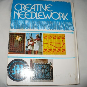 May include: A book titled "Creative Needlework" with a blue cover and a white border. The cover features a rug, two women, a woven pattern, and a logo with the letter "M".
