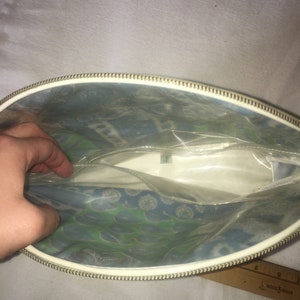 Op de afbeelding: Een witte vinyl toilettas met een gouden rits. De tas heeft een transparante voorzijde van plastic met een blauw en groen bloemenpatroon.