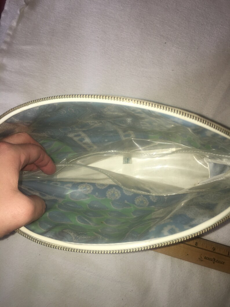 Op de afbeelding: Een witte vinyl toilettas met een gouden rits. De tas heeft een transparante voorzijde van plastic met een blauw en groen bloemenpatroon.
