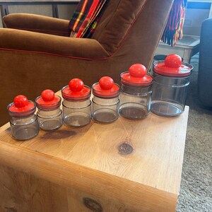May include: A set of six clear glass jars with bright red lids, arranged in a row on a wooden surface. The jars vary in size, with the smallest on the left and the largest on the right. The lids are round and have a knob on top.