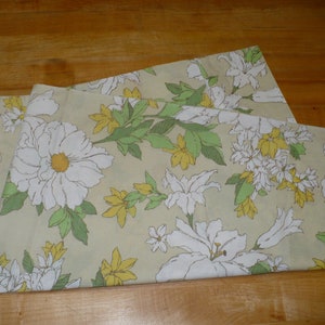 Puede incluir: Una tela doblada con un patr&oacute;n floral. La tela presenta flores blancas con centros amarillos, hojas verdes y un fondo beige claro.