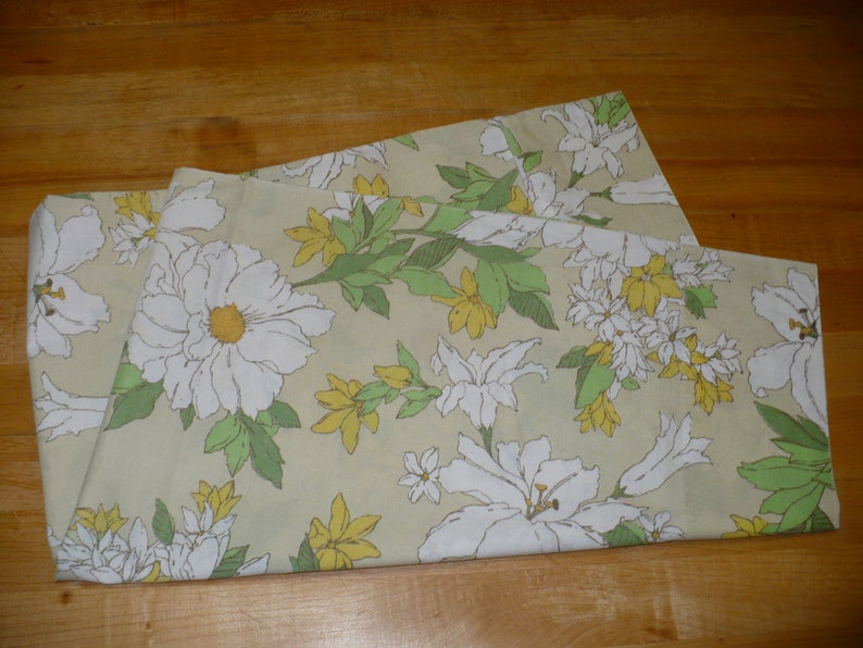 Puede incluir: Una tela doblada con un patr&oacute;n floral. La tela presenta flores blancas con centros amarillos, hojas verdes y un fondo beige claro.