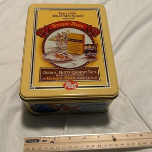 May include: A rectangular yellow metal tin featuring vintage Grape-Nuts cereal branding, including the text Every table should have its daily ration of Grape-Nuts, Original Nutty Crunchy Taste, and Post. A ruler shows the tin is 8 inches wide.
