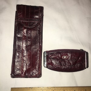May include: Two burgundy leather wallets with a silver zipper and a silver clasp. The larger wallet has a flap closure. The smaller wallet has a zipper closure and a silver logo with the letters "C" and "V" intertwined.