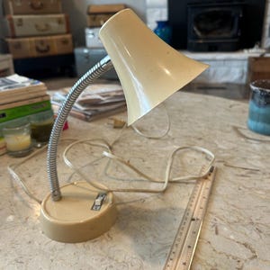 May include: A vintage beige desk lamp with a flexible, silver-colored neck. The lamp has a round base and a flared lampshade. A wooden ruler is next to the lamp. The lamp is on a marble-patterned surface.