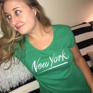 May include: A woman wearing a green t-shirt with the words "New York" written in white on the front.