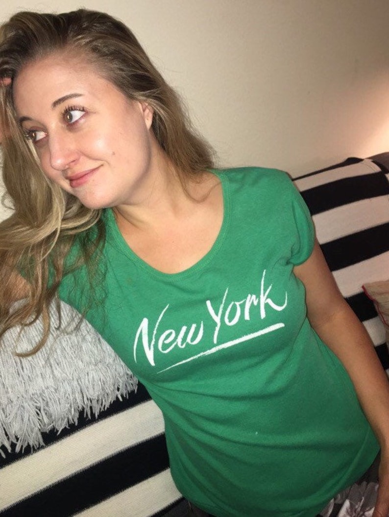 May include: A woman wearing a green t-shirt with the words "New York" written in white on the front.