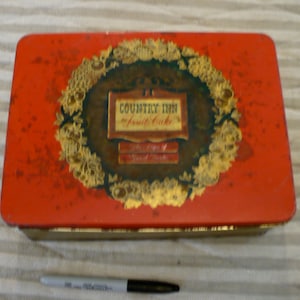 May include: Red metal tin with a gold trim and a green and gold wreath design. The tin has a label that reads "Country Inn Fruit Cake".