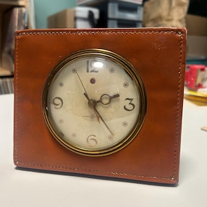 May include: A square analog desk clock encased in brown leather with visible stitching along the edges. The clock face is off-white with a brass-toned circular frame, featuring black numerals at the 3, 6, 9, and 12 positions, and a small General Electric logo.