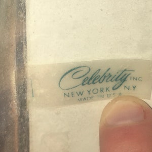 Op de afbeelding: Een wit label met de tekst "Celebrity Inc. New York, NY Made in USA" in turquoise letters.